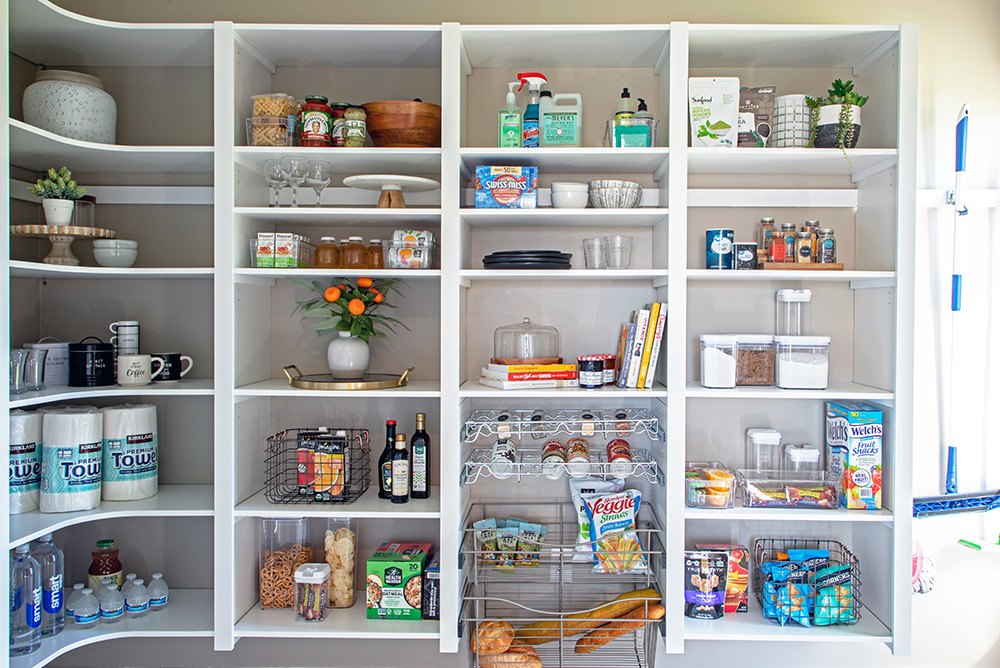Pantry wall in white with rounded corner on left. Food item storage on shelves and glide-out wire baskets with cans, snacks and bread.